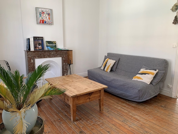 Een woonkamer met een houten salontafel, een grijze bank en een open haard met boeken erboven.