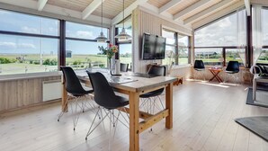 Een ruime eethoek met een houten tafel en stoelen, grote ramen met uitzicht op het platteland, en een aan de muur bevestigde televisie.