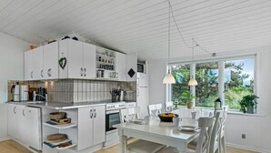 A modern kitchen with white cabinetry, a dining area with a wooden table and chairs, and a large window offering a view of greenery. - "Stina" - 250m from the sea by Interhome (Løkken)