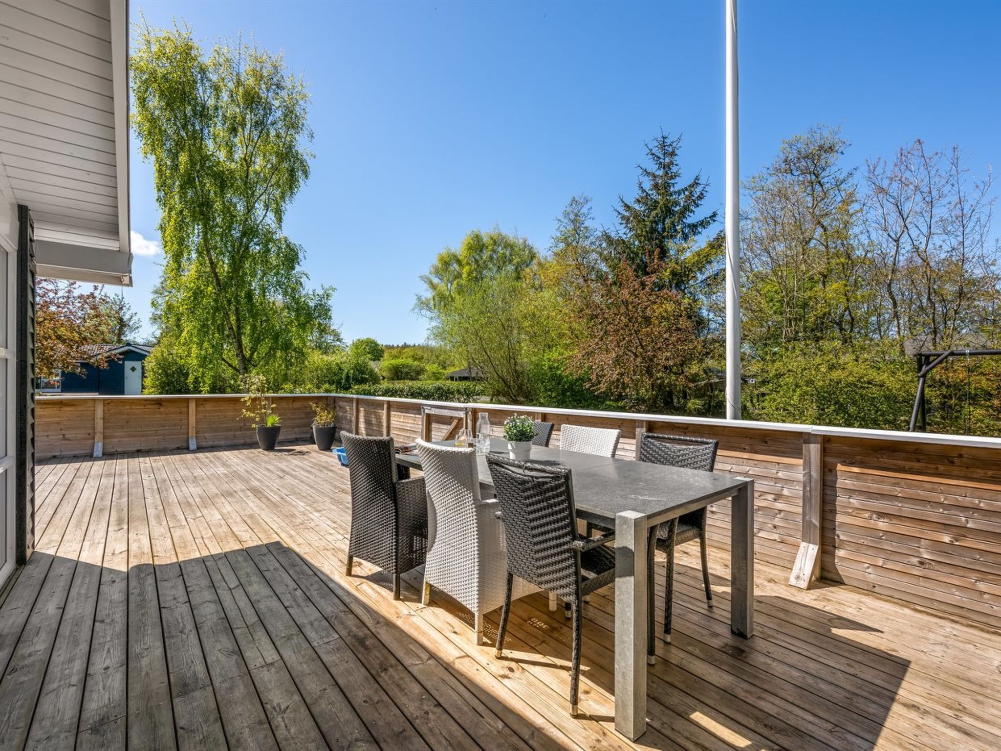 Eine Holzterrasse mit Gartenmöbeln, darunter ein Tisch und Stühle, umgeben von Bäumen und blauem Himmel.
