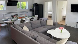 Ein modernes Wohnzimmer mit grauem Sofa, einem Essbereich mit Tisch für zwei Personen und einer Küche mit schwarzem Schranksystem sowie einem an der Wand montierten Fernseher.