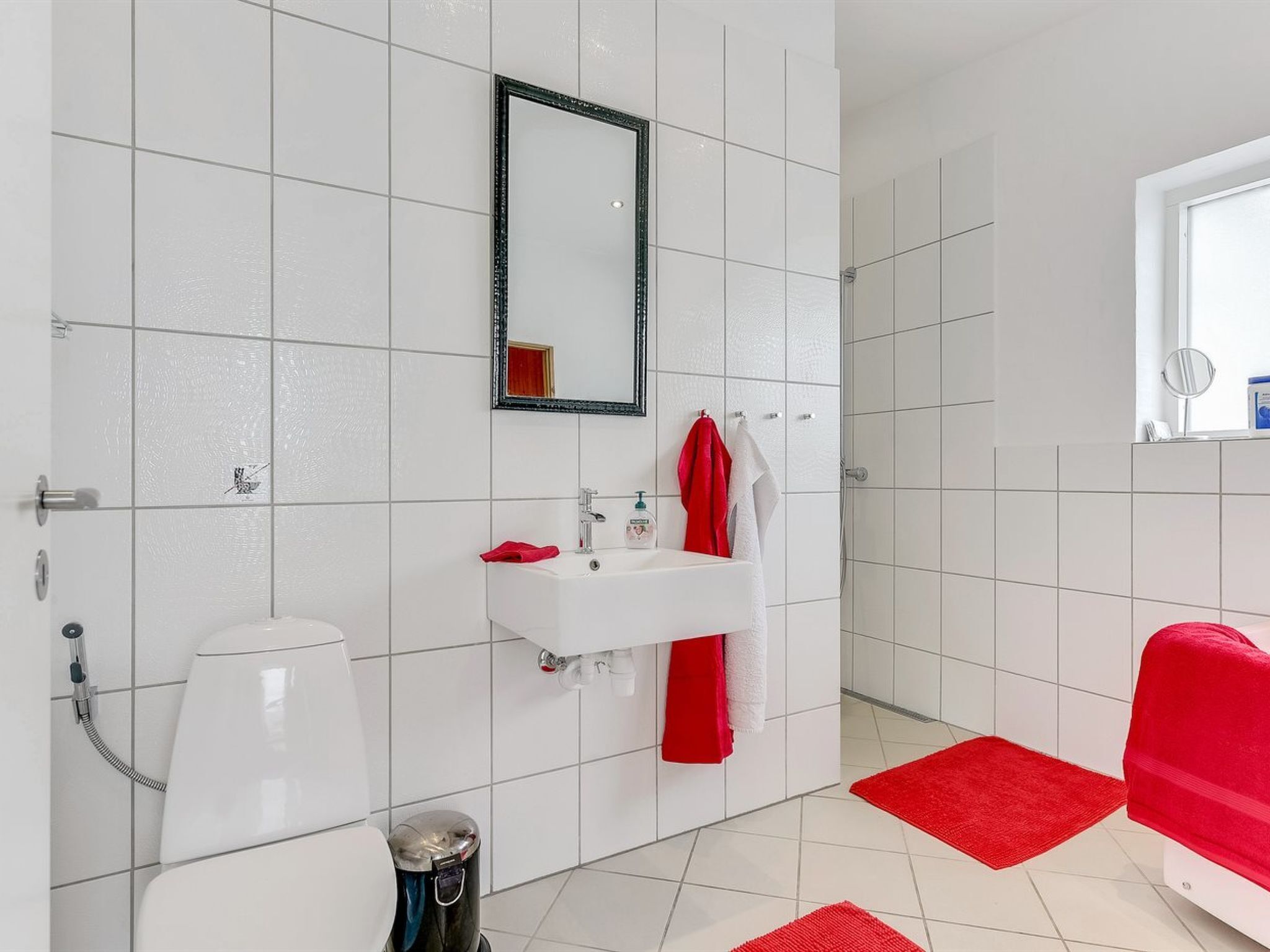 Une salle de bain avec des carreaux blancs, un lavabo blanc, un miroir, une serviette rouge et un tapis de bain rouge.