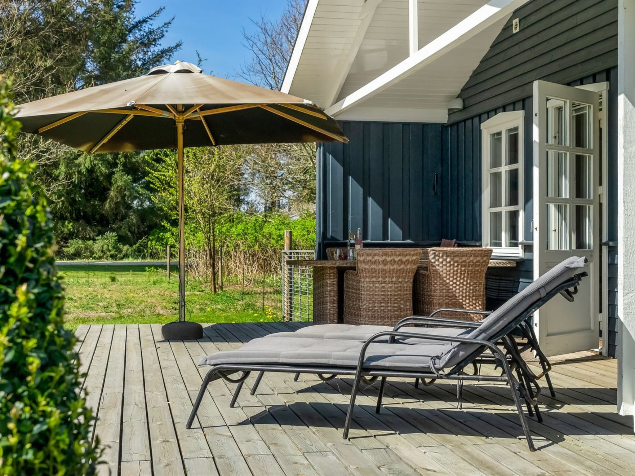 An outdoor deck with two lounge chairs and a large umbrella, a wicker table, and a barbecue grill.