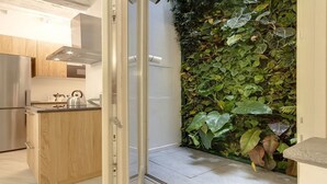 Uma cozinha moderna com geladeira de aço inoxidável, armários de madeira e bancada com chaleira. Uma porta de vidro deslizante dá acesso a uma varanda com parede verde exuberante de plantas.