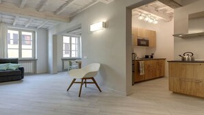Uma sala de estar moderna com um sofá e uma poltrona, área de cozinha com armários de madeira e uma chaleira, e grandes janelas que permitem entrada de luz natural.