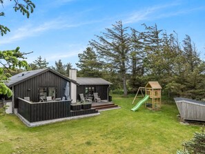 A wooden cabin with a deck and outdoor seating, surrounded by a grassy yard and trees.