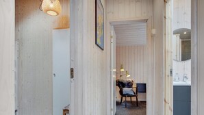 Ein schmaler Korridor mit Holzwänden, einer Bank und einer Tür zu einem Zimmer mit Bett und Schreibtisch.
