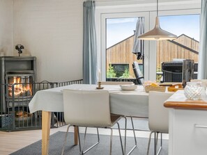 A cozy room with a fireplace, a dining table with chairs, a hanging pendant light, and a view of a wooden building outside.