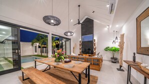 Un espace repas moderne avec une table et des bancs en bois, un ventilateur de plafond, des luminaires suspendus et un grand écran de télévision.