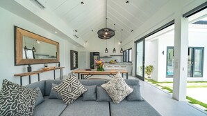 Un salon moderne avec un canapé d’angle gris, des coussins à motifs, une table basse en bois et des luminaires suspendus.