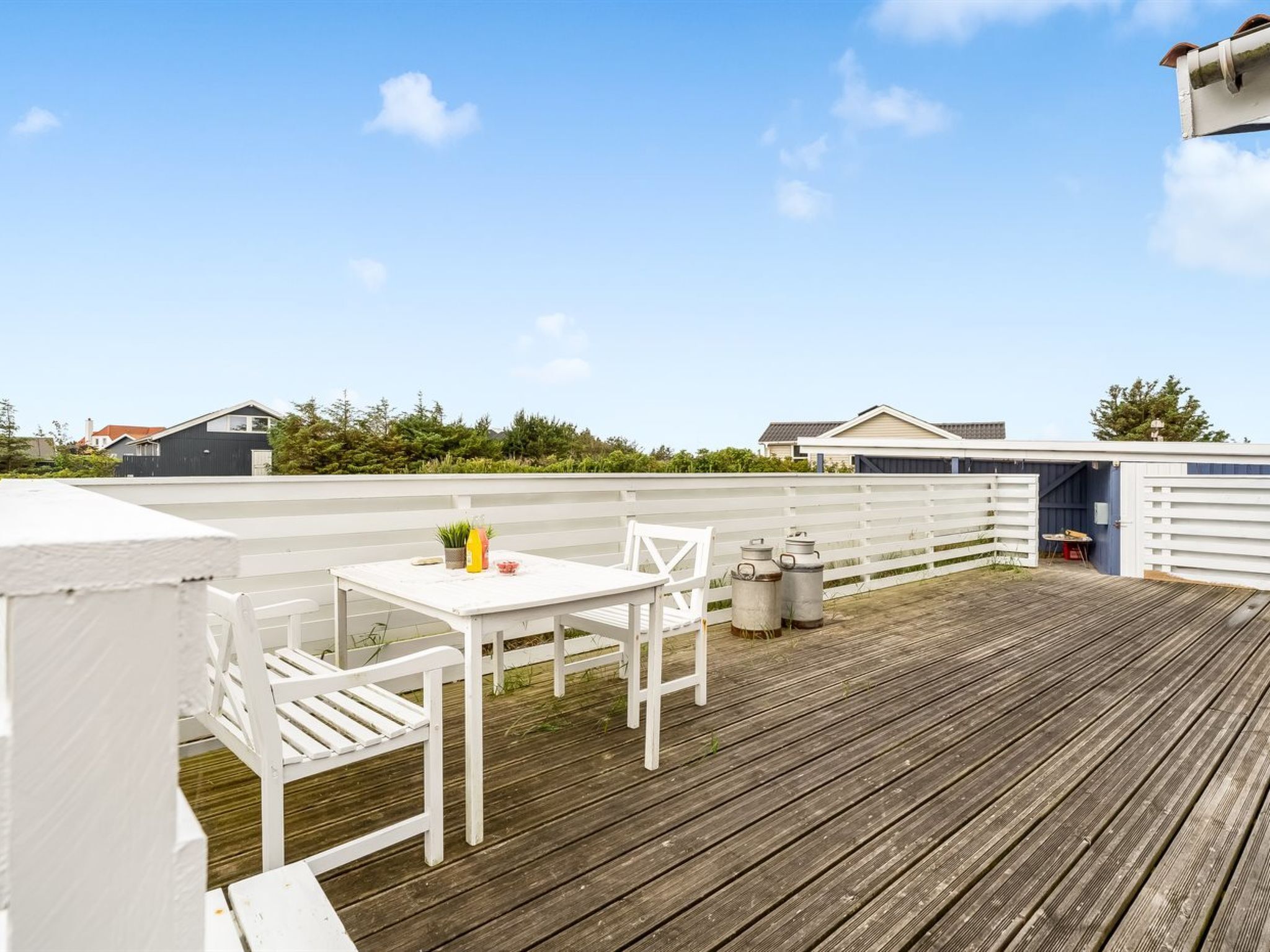 A wooden deck with a white table and chairs, a small table with a plant and a bottle, and a covered area with a door.