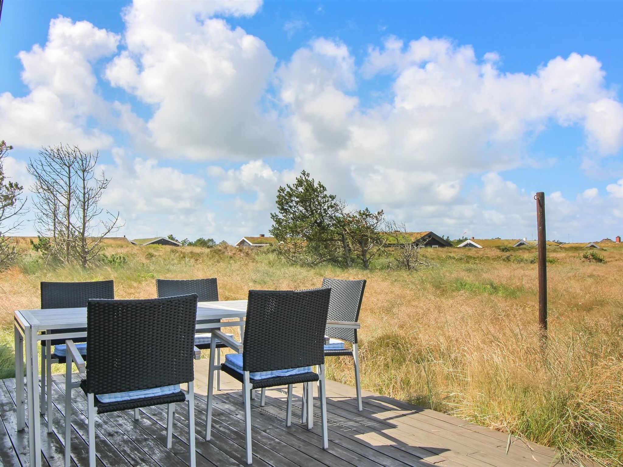 A wooden deck with a table and four chairs, overlooking a grassy field with houses in the distance.