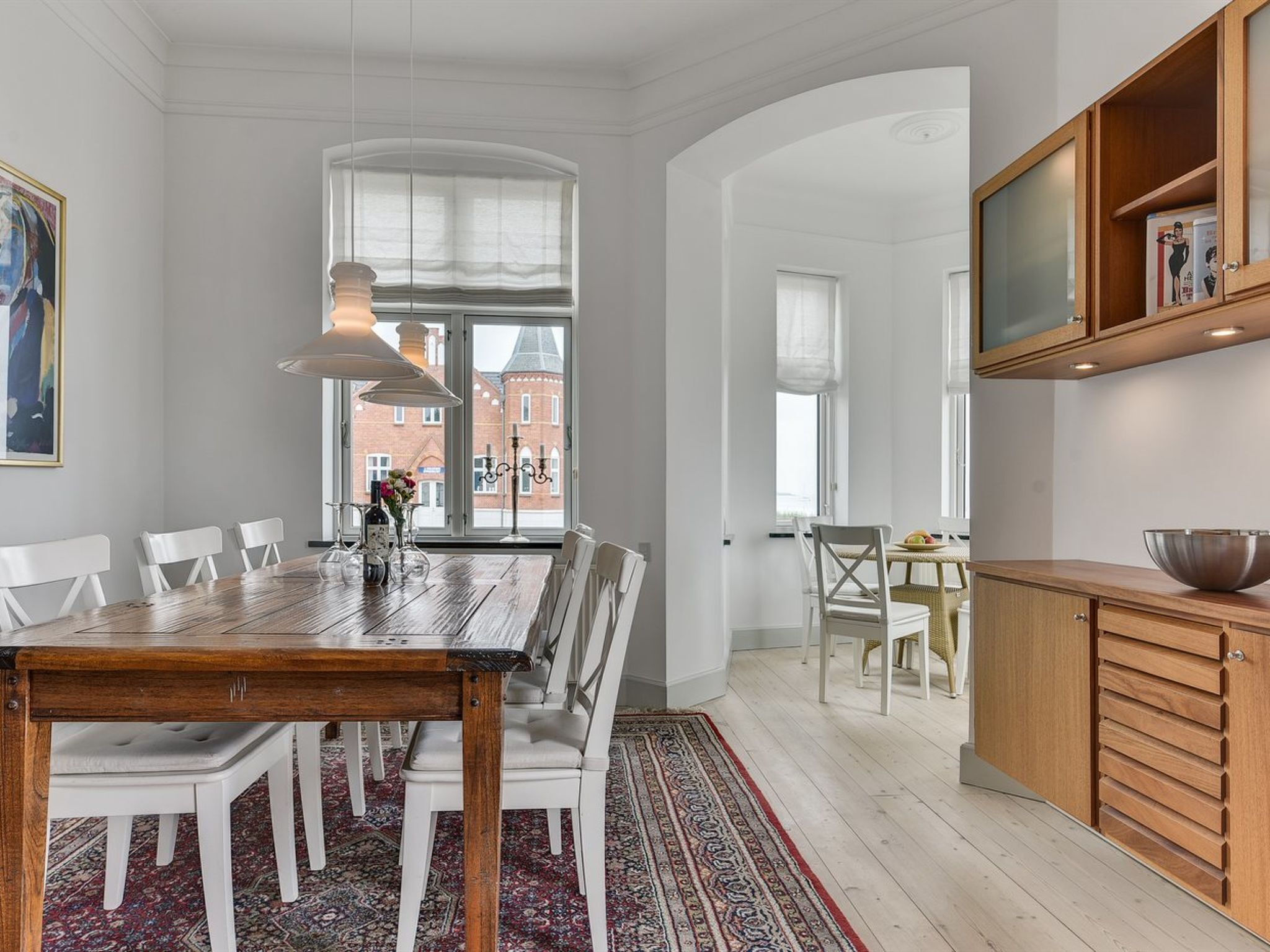 A dining area with a wooden table and white chairs, a patterned rug, and a view of a brick building through large windows.