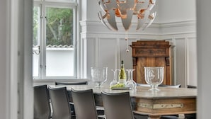 Une salle à manger avec une table en bois, des chaises noires, un lustre et une vue sur l’extérieur par une fenêtre.