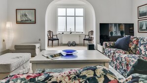 Un salon avec un canapé, une table basse et un téléviseur. Il y a une fenêtre donnant sur la mer et une chaise près de la fenêtre.