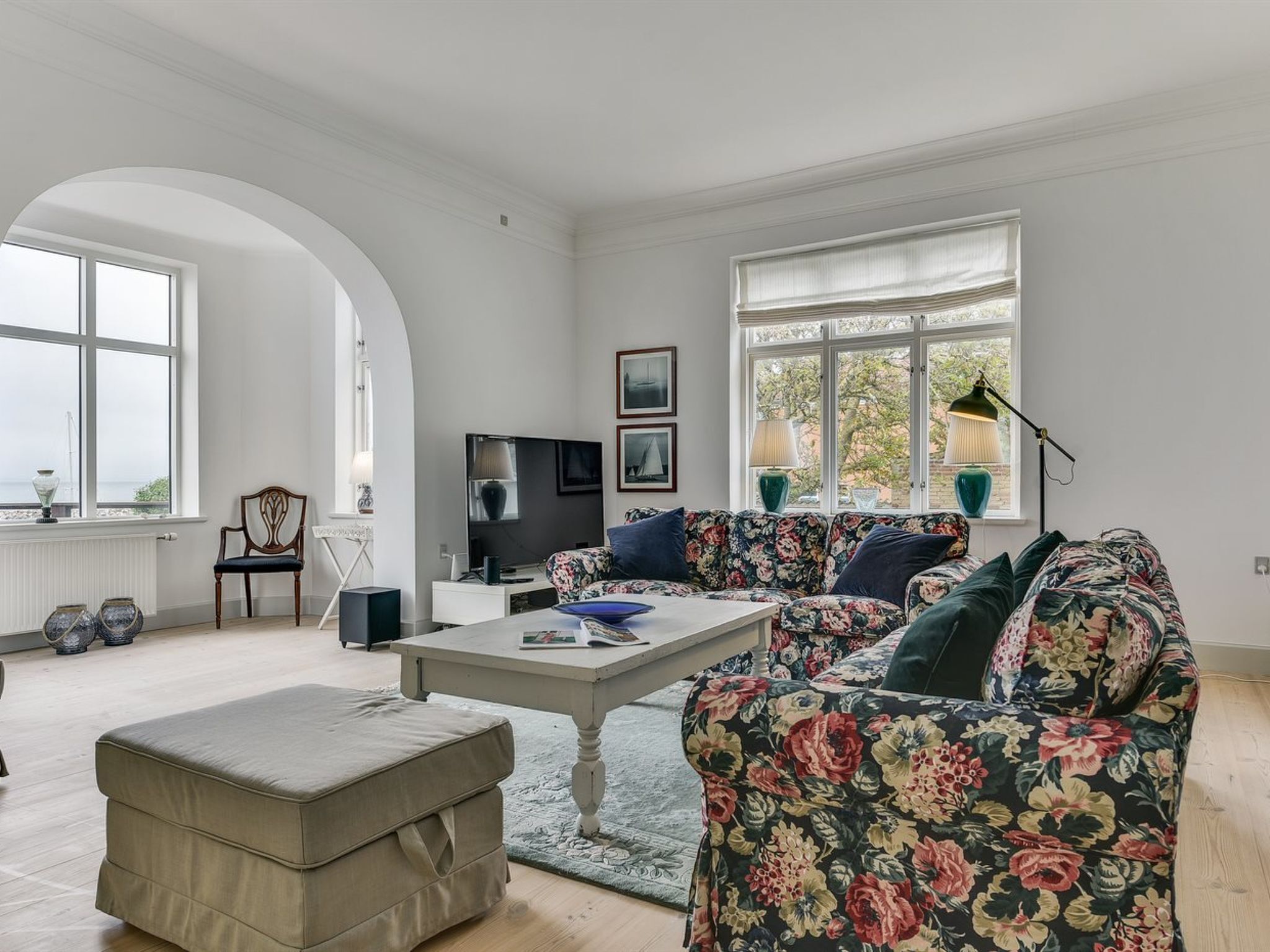 Un salón espacioso con un sofá grande estampado con flores, una mesa de centro y un televisor. Hay dos ventanas que dejan entrar luz natural y ofrecen vistas al exterior.