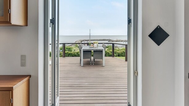 Une porte ouverte donne sur une terrasse où se trouvent une table et des chaises, d’où l’on peut admirer un plan d’eau et une côte rocheuse.