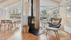 Un intérieur moderne avec un poêle à bois noir, un plancher en bois, un coin repas avec une table et des chaises blanches, et un coin salon confortable avec un fauteuil gris et un panier en osier.