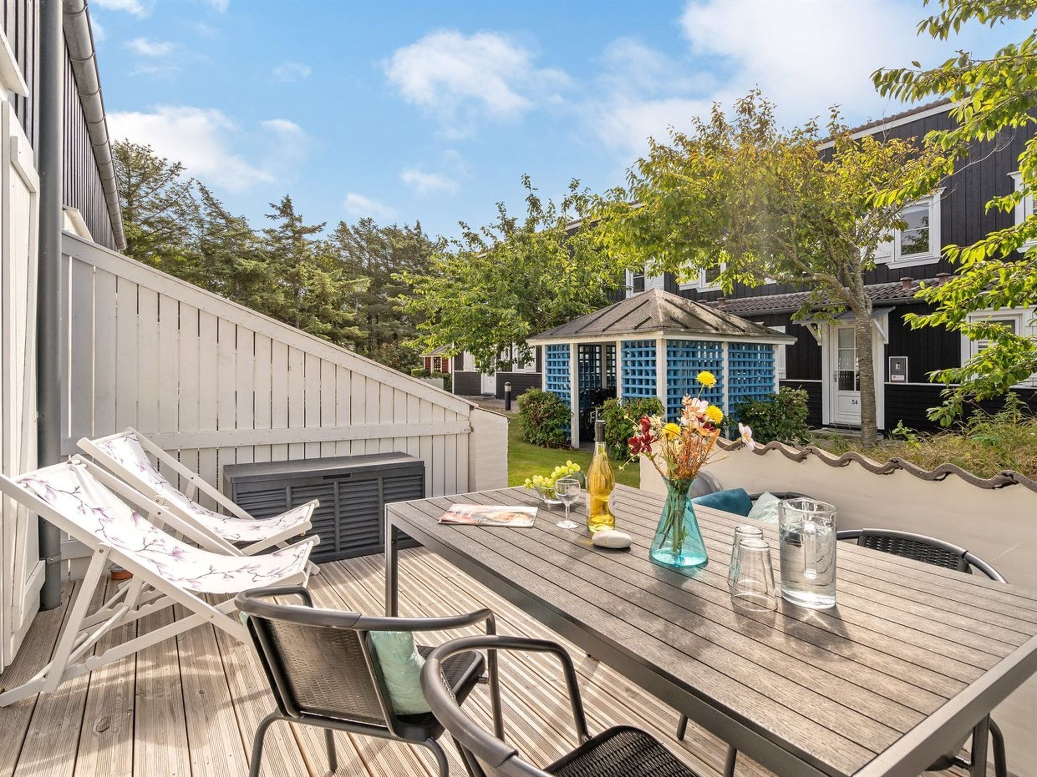 A wooden deck with a table and chairs, a gazebo, and a garden with trees and a fence.