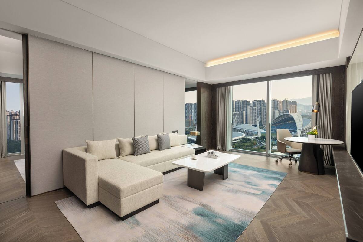 Un salon moderne avec un canapé d’angle, une table basse et une vue sur le paysage urbain grâce à de grandes fenêtres.