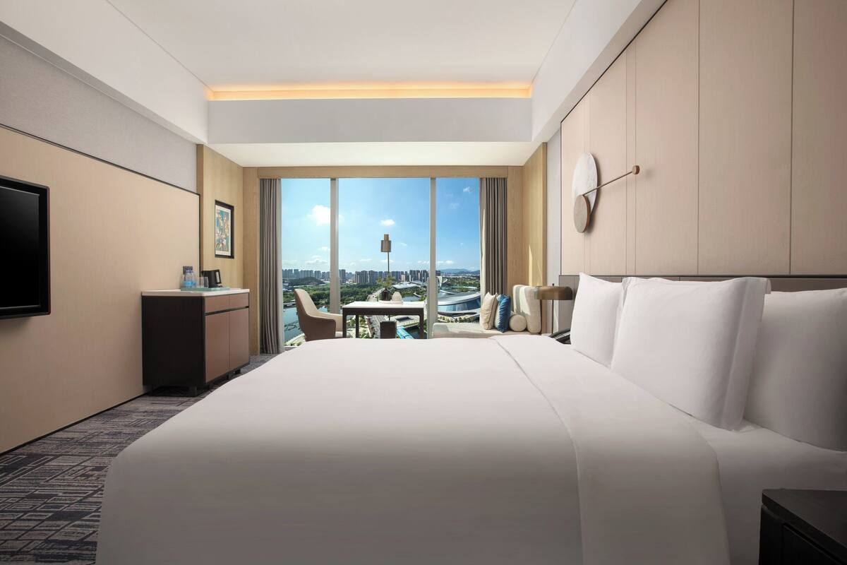 Une chambre d’hôtel avec un grand lit, un bureau, une chaise et une télévision. Une grande fenêtre offre une vue sur les bâtiments et le paysage urbain.