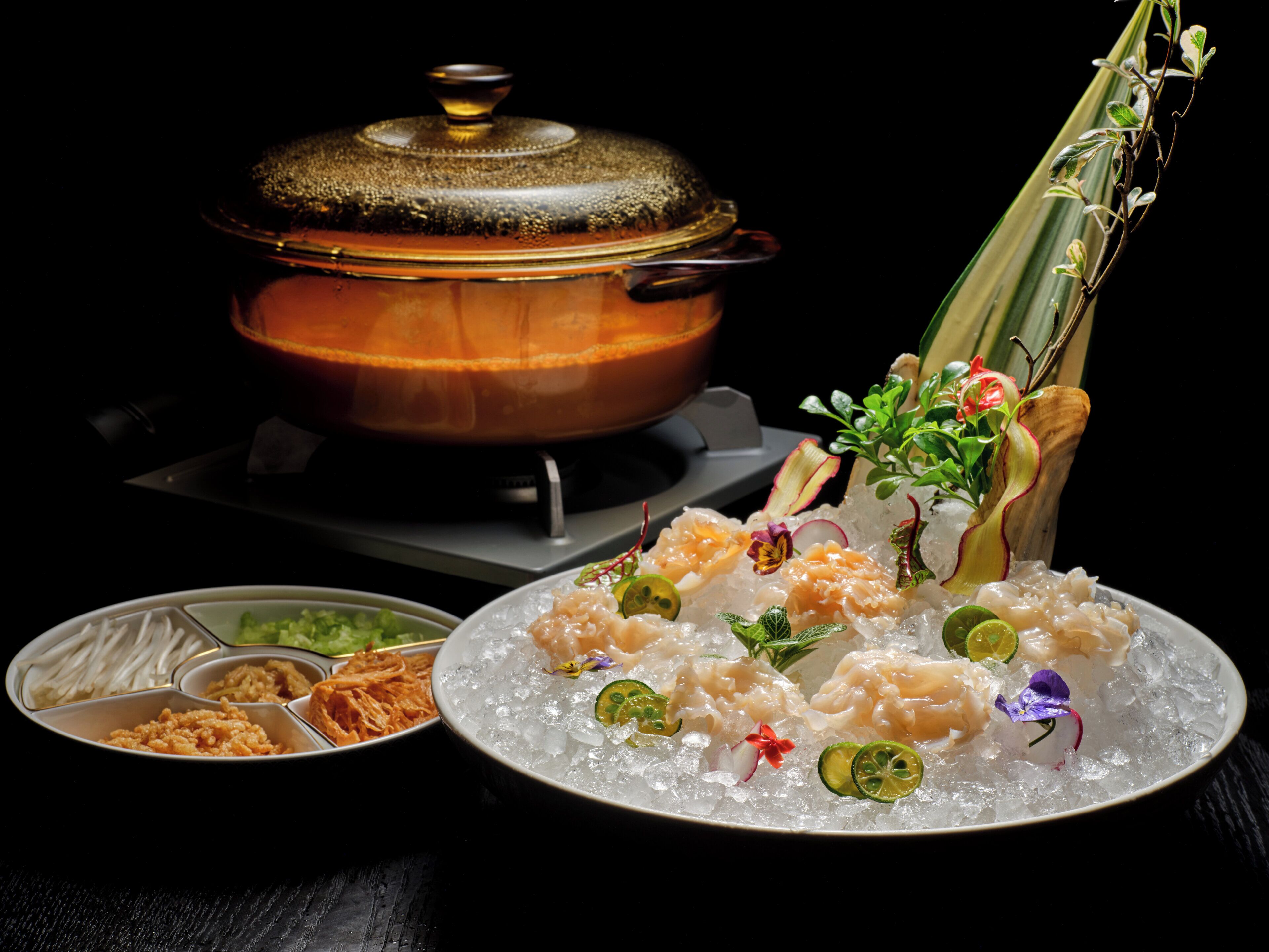A dining setup with a pot on a stove, a plate with seafood and garnishes, and a side dish with vegetables and sauces.