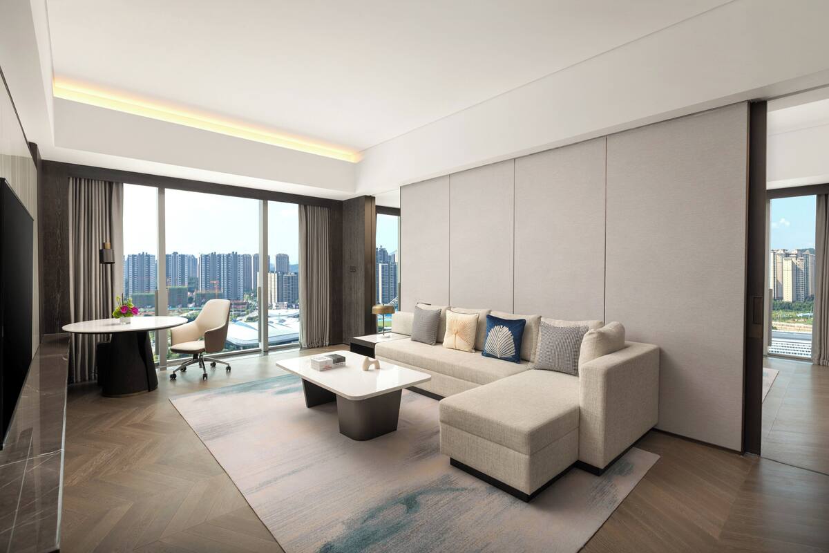 Un salon moderne avec un canapé d’angle, une table basse et un coin repas comprenant une table et des chaises. De grandes fenêtres offrent une vue sur le paysage urbain.