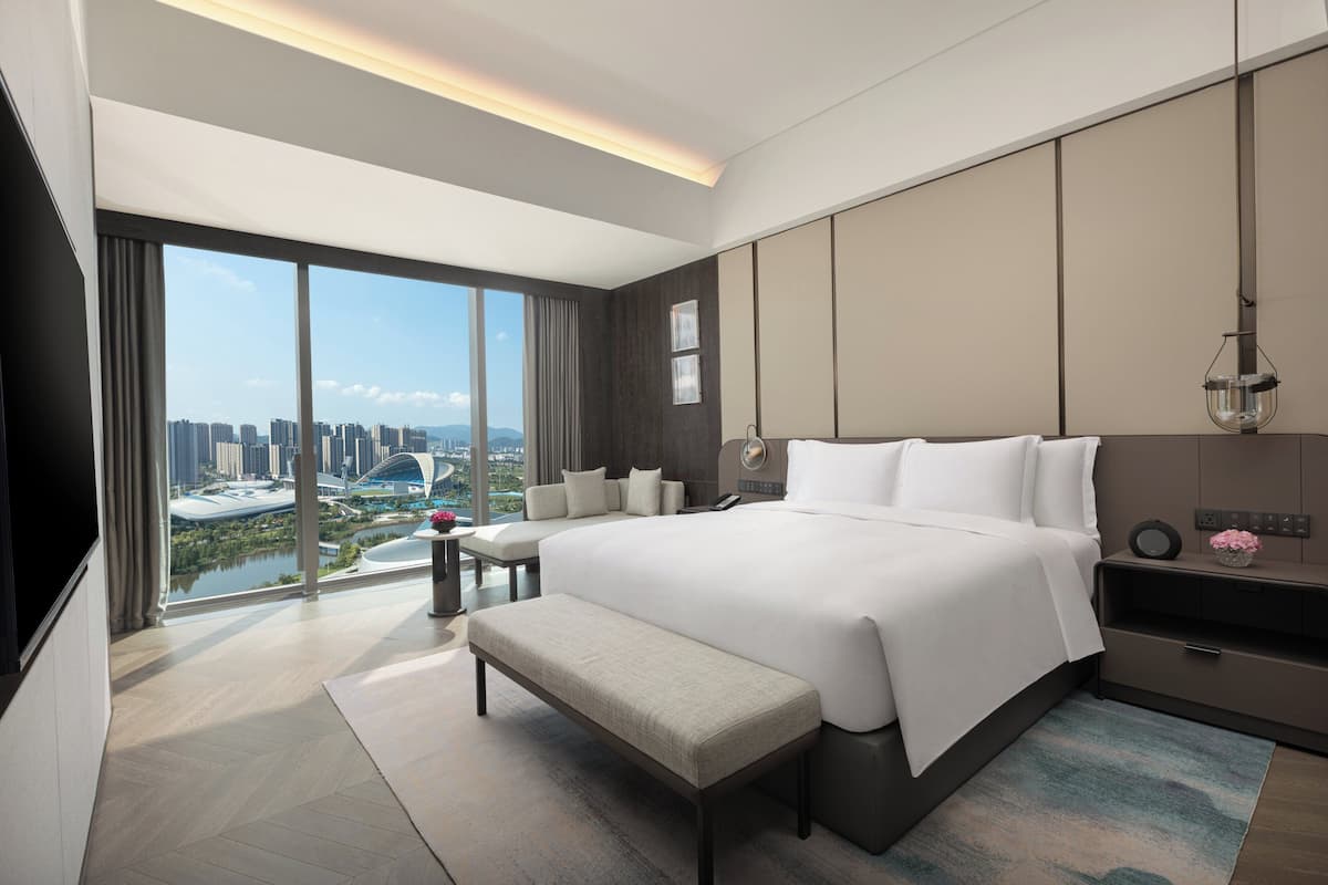 Une chambre d’hôtel moderne avec un grand lit, une banquette, une table de chevet et une vue sur le paysage urbain.