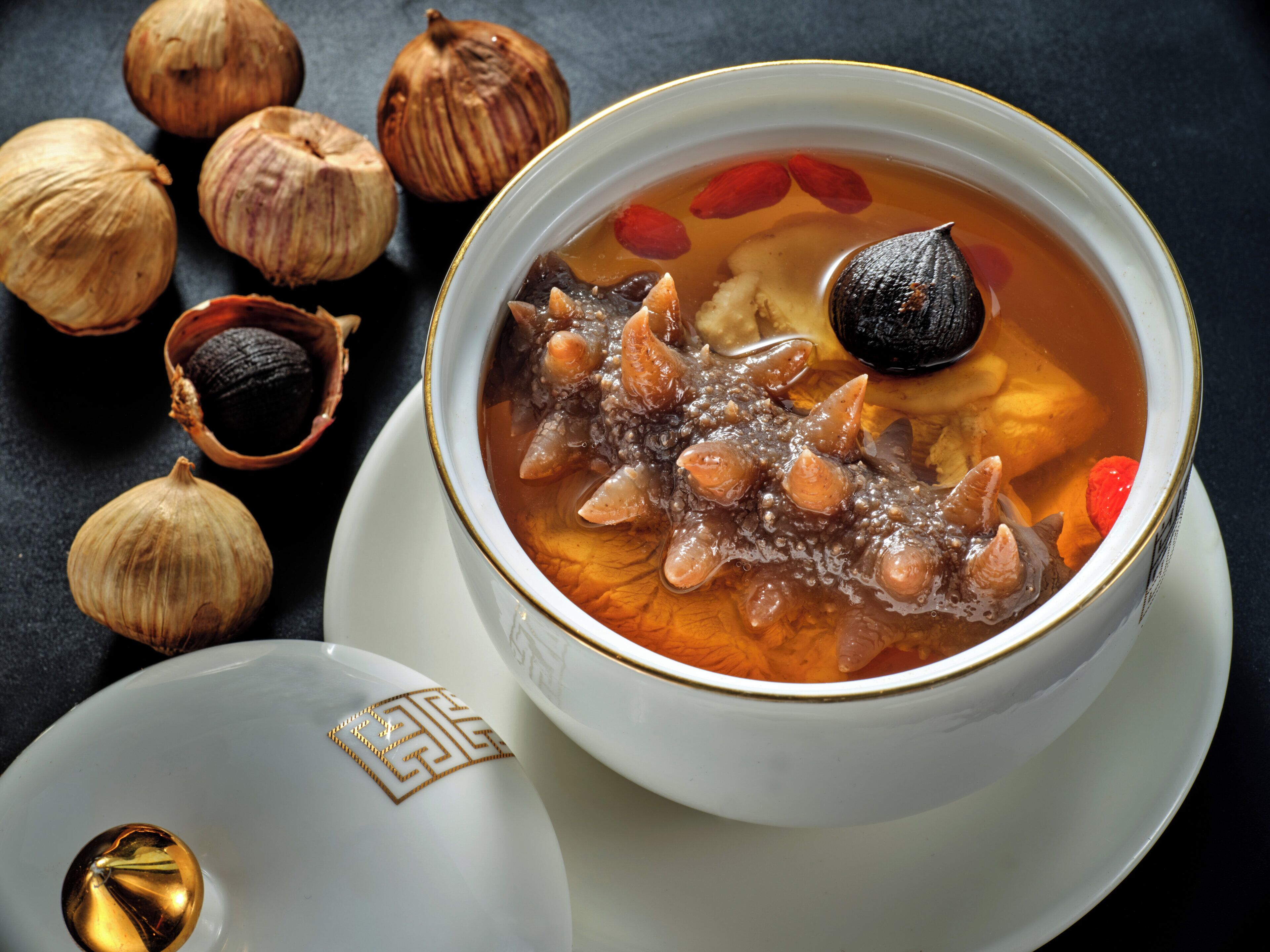 Un tazón de sopa con diversos ingredientes, acompañado de dientes de ajo enteros y una cuchara decorativa.