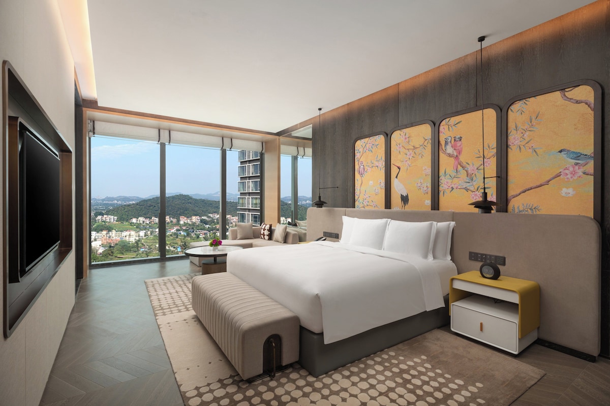 A modern hotel room with a large bed, a TV, a nightstand, and a view of the city.