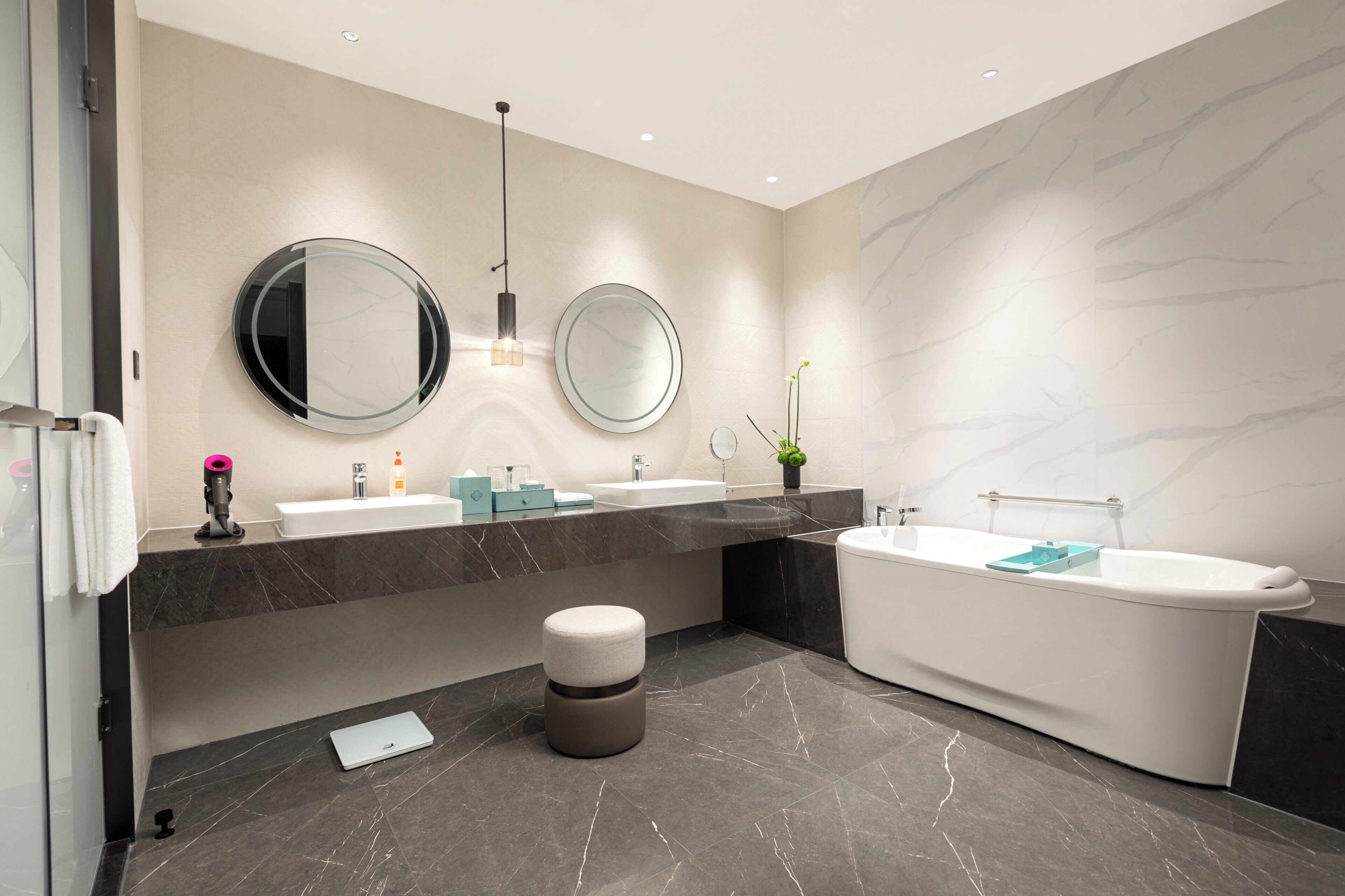 A modern bathroom with a large bathtub, two sinks, a round mirror, and a small stool.