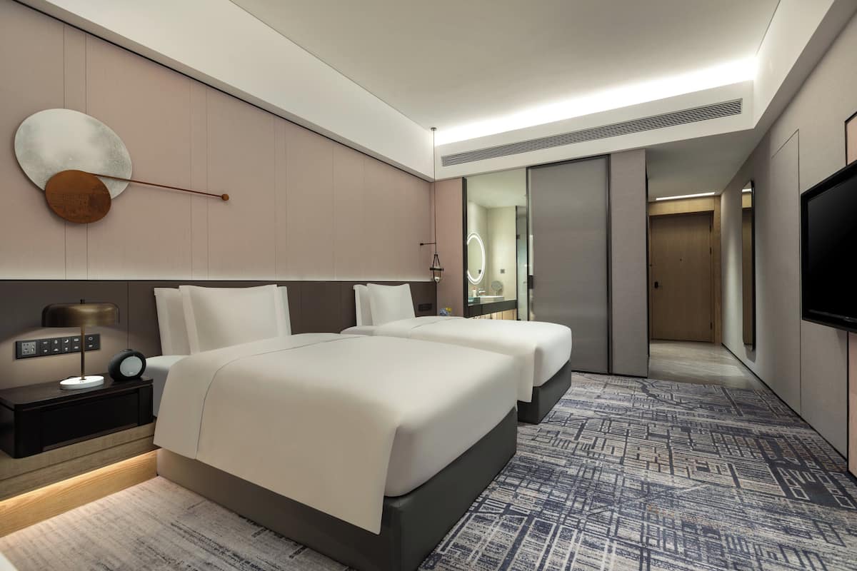 Une chambre d’hôtel moderne équipée de deux lits, d’une table de chevet avec une lampe, d’un grand miroir et d’un téléviseur à écran plat.
