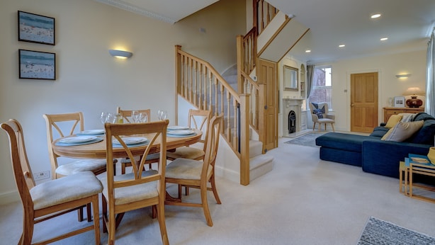 A dining area with a wooden table and chairs, a staircase, and a living room with a blue sofa. - The Old Village Shop | Spacious Dog Friendly Cottage Near the Beach (Winterton-on-Sea)