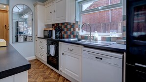 A modern kitchen with white cabinetry, a herringbone wood floor, a large window with a view of a brick building, and a dishwasher. - The Old Village Shop | Spacious Dog Friendly Cottage Near the Beach (Winterton-on-Sea)