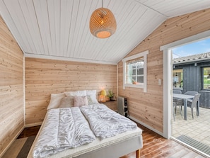 A room with wooden walls and ceiling, a bed with white bedding, a hanging light fixture, and a window overlooking an outdoor area.