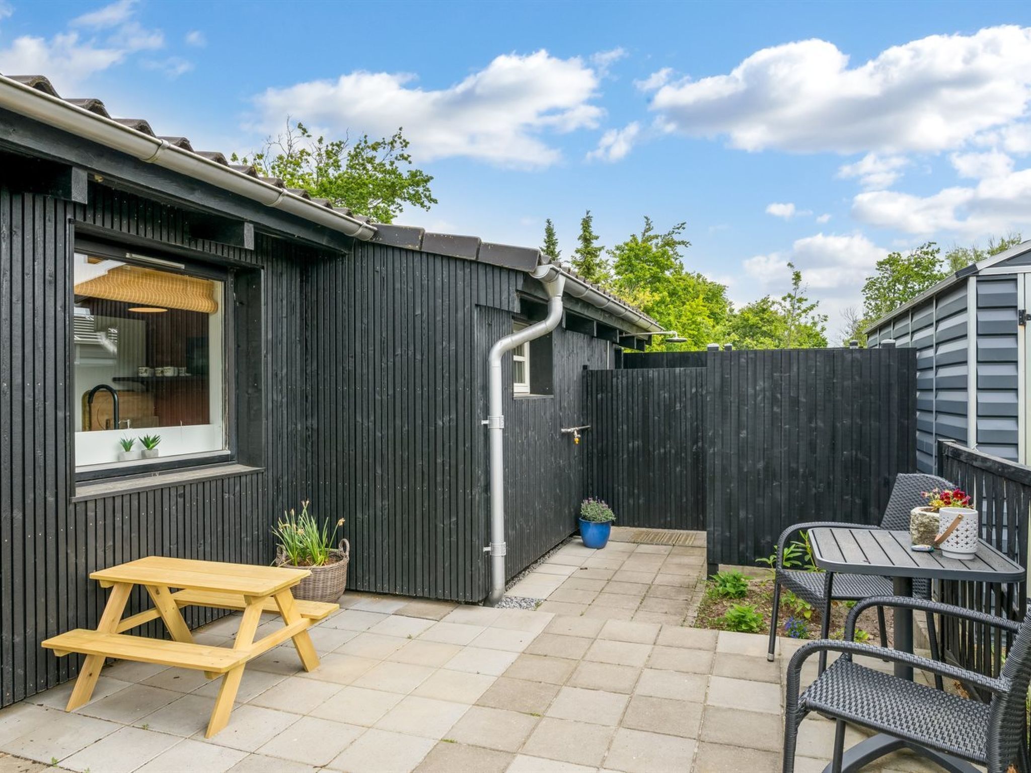 A patio with a wooden table and chairs, a small garden area with potted plants, and a building with a window and a door.