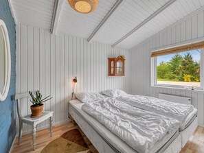 Ein Schlafzimmer mit einem Bett, einem kleinen Tisch mit Pflanze, einer Wandlampe, einem Fenster mit Blick auf Bäume und einem Holzschrank.