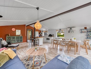 A spacious living area with a sofa, coffee table, and dining table. There is a kitchen with a dining area, a brick wall, and a large window.
