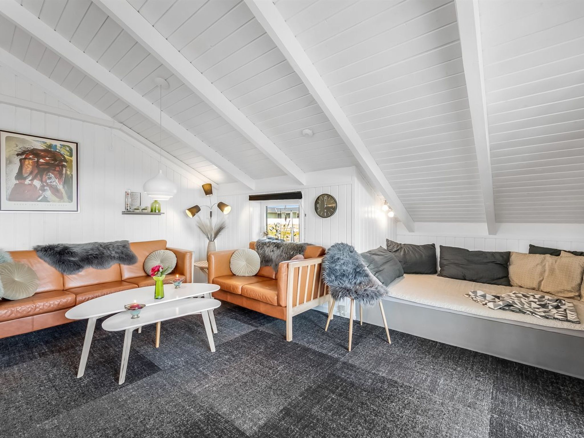 A modern living room with a high ceiling, white walls, and a mix of leather and fabric sofas. There is a white coffee table, a floor lamp, and a clock on the wall.