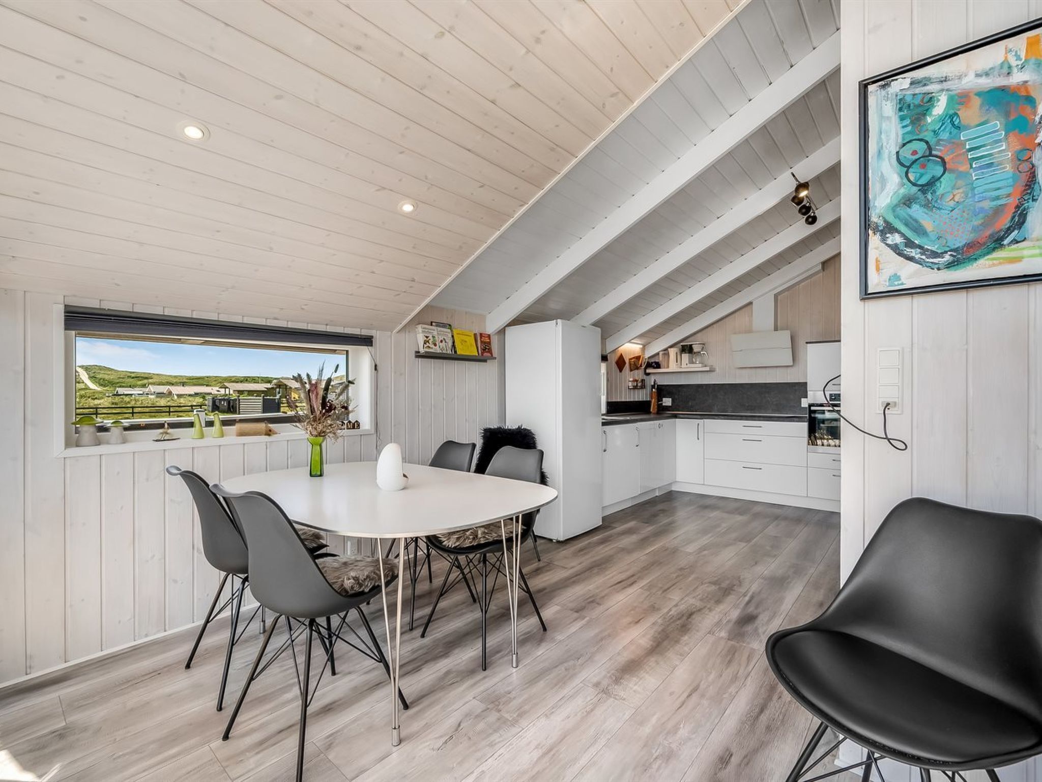 Une cuisine moderne avec des meubles blancs, un coin repas doté d’une table et de chaises blanches, et une vue sur la campagne.