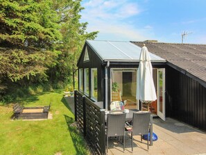 Une petite maison de couleur sombre, dotée d’une porte en verre qui donne sur un patio. On y trouve une table avec des chaises et un parasol blanc à l’extérieur.