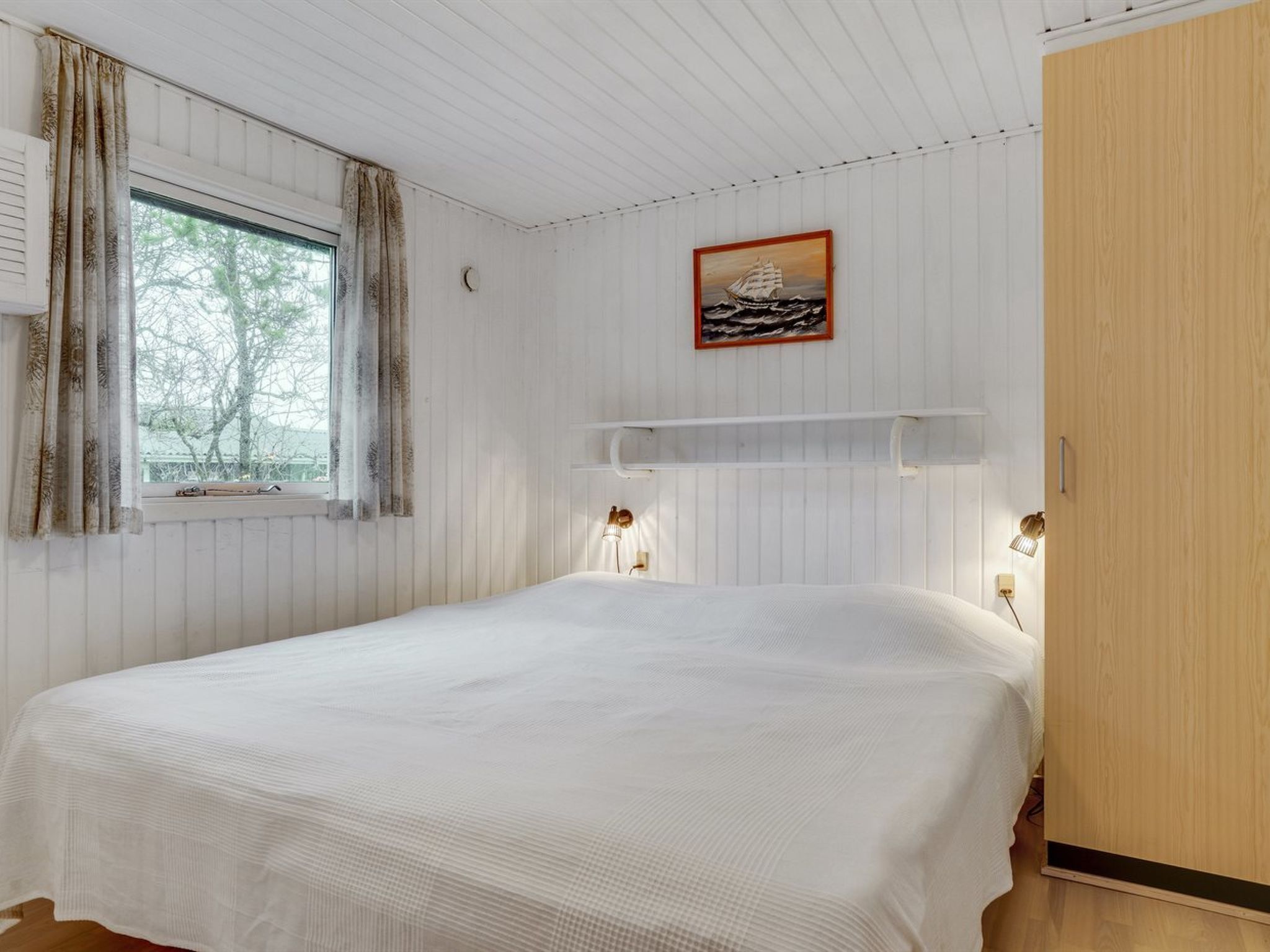 Une chambre à coucher avec un lit, une fenêtre avec des rideaux, une étagère fixée au mur avec un tableau, et une table de chevet avec une lampe.
