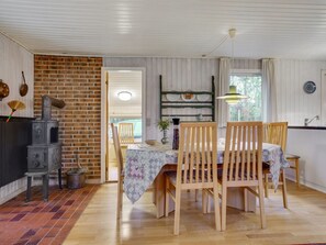 Un coin repas chaleureux avec un mur en briques, des chaises en bois, une table recouverte d’une nappe à motifs floraux et un poêle d’époque.