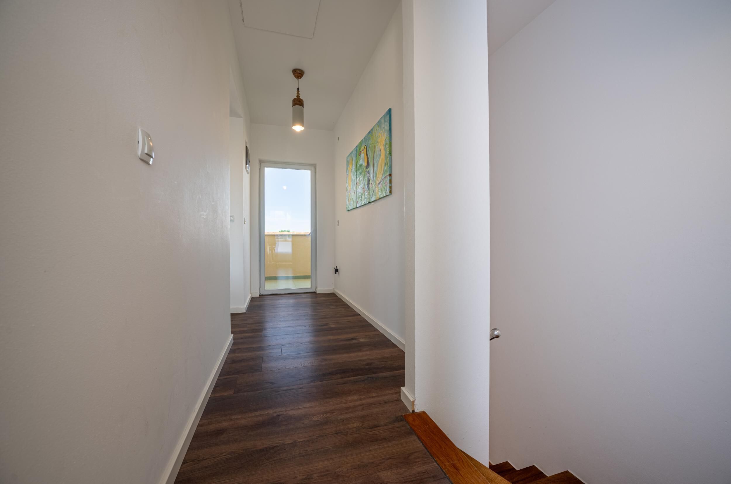 Een hal met houten vloer, witte muren, een plafondlamp en een deur naar een balkon.