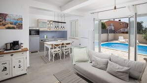Ett modernt kök med vita skåp, en matplats med bord och stolar samt ett stort fönster med utsikt över en swimmingpool.