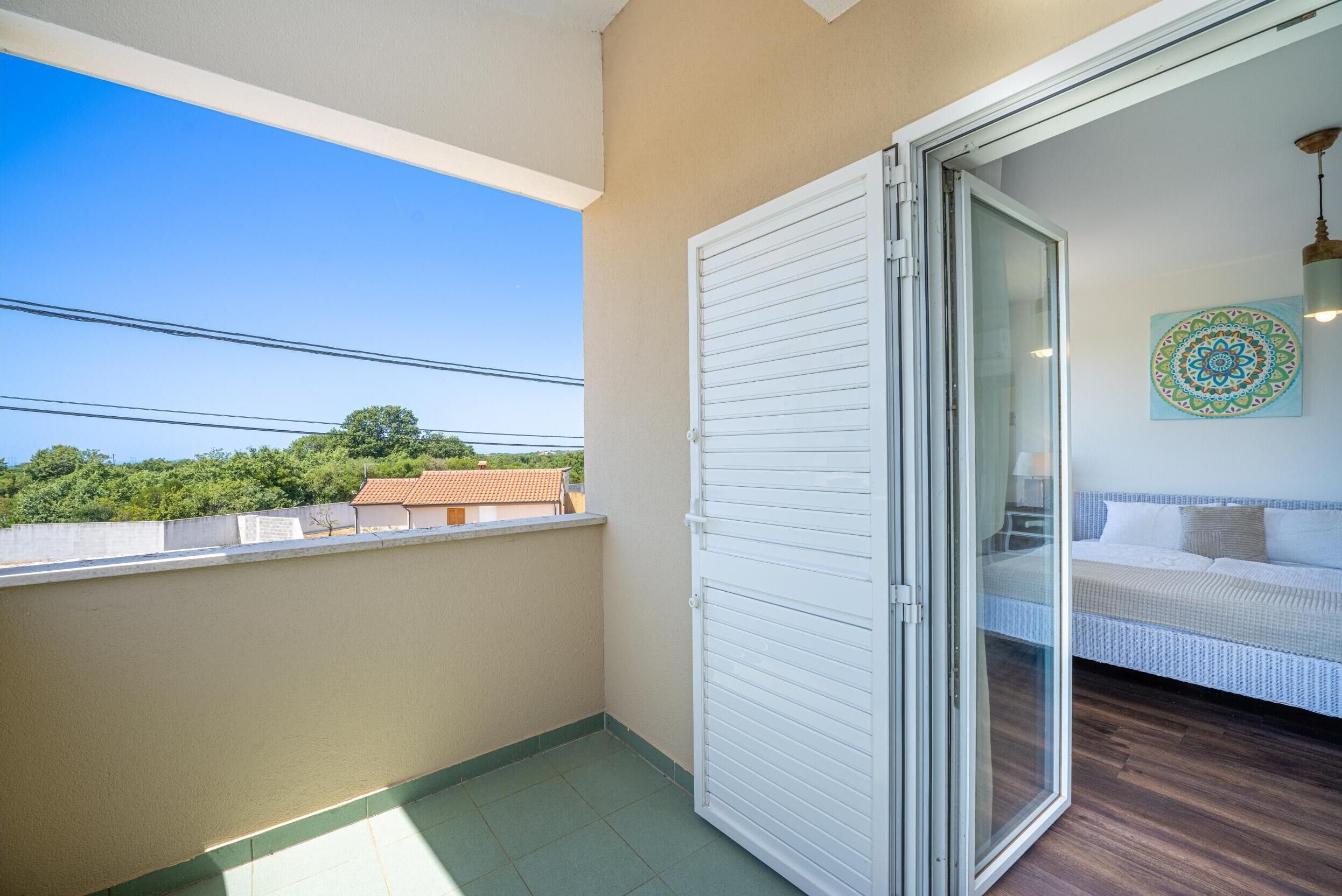 A balcony with a view of trees and buildings, a sliding glass door leading to a bedroom with a bed and a decorative wall piece.