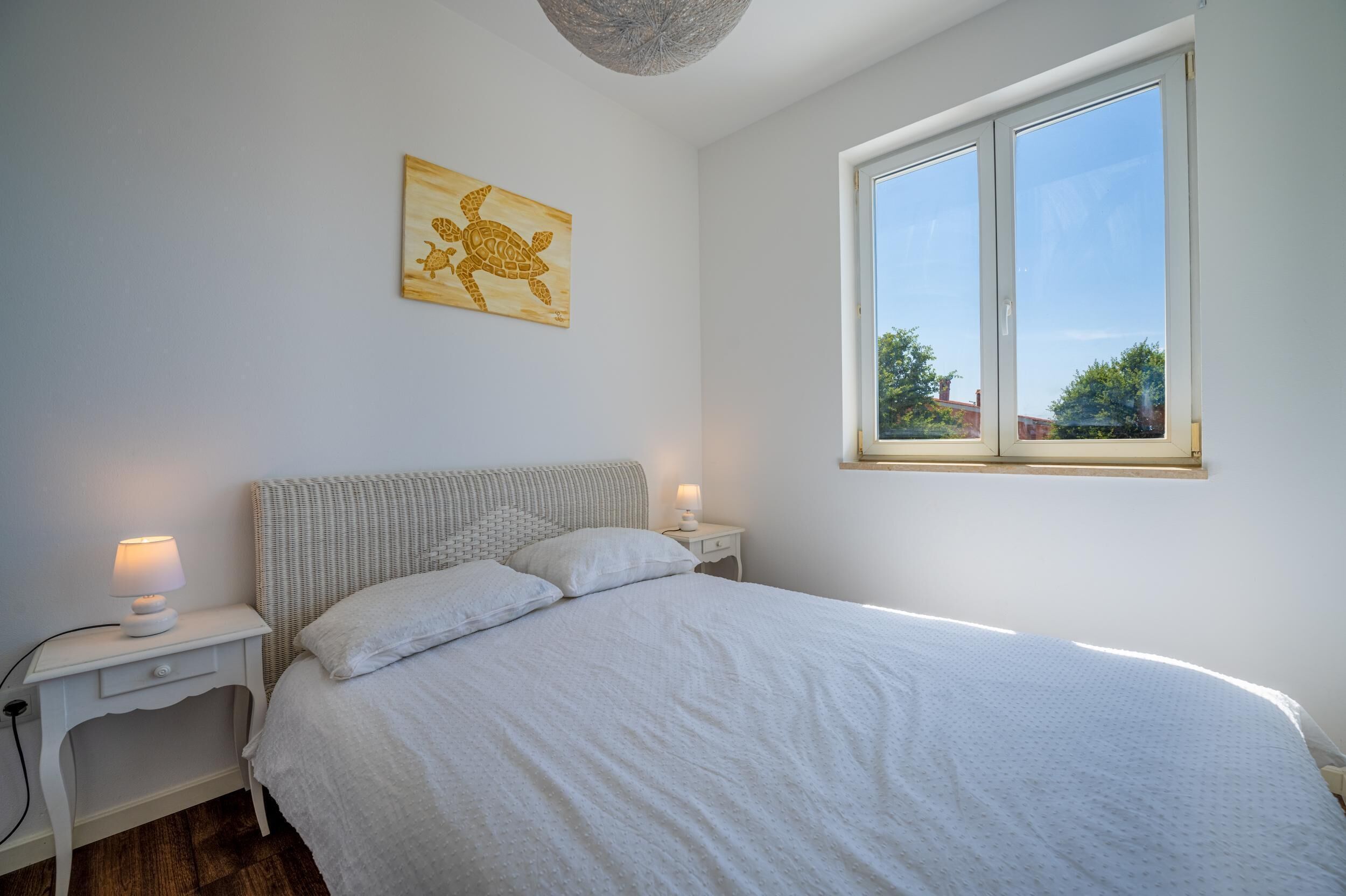 Un dormitorio con una cama grande, dos mesitas de noche con lámparas, un cuadro de pared de madera con una tortuga y una ventana con vistas a árboles y edificios.
