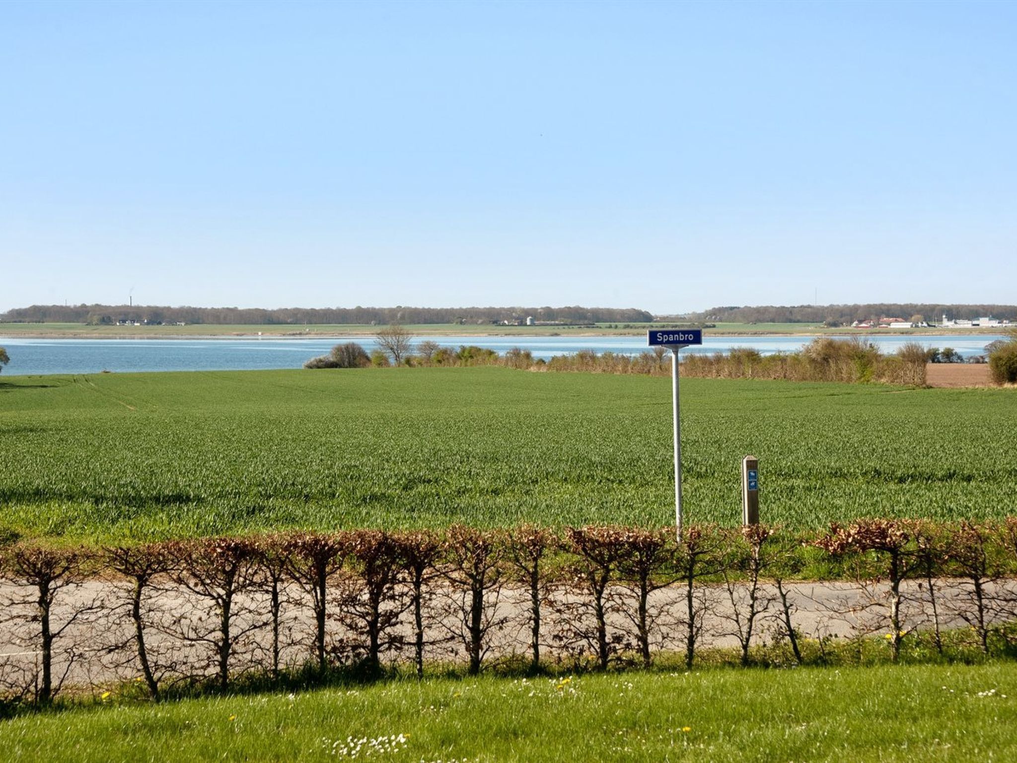 Ländliche Landschaft mit einem Schild, auf dem 'Spanbro' steht, einem Feld, einem Gewässer und einer fernen Baumgrenze.