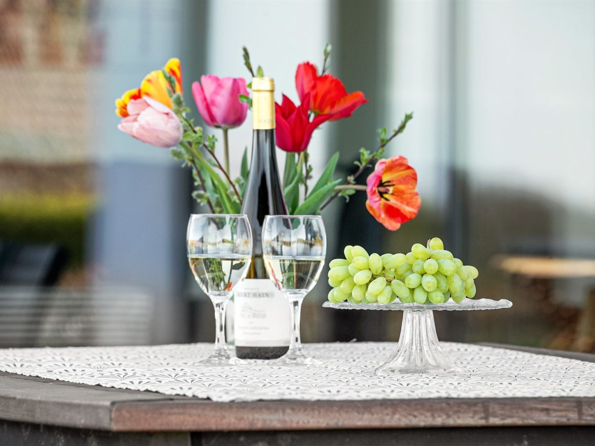 Ein Tisch mit einer Weinflasche, zwei Gläsern, einer Traubenteller und bunten Blumen.