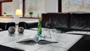 A modern living room with a leather sofa, a coffee table with a book, glasses, and a Sprite can. There are two windows with views of trees and a lamp on a side table.